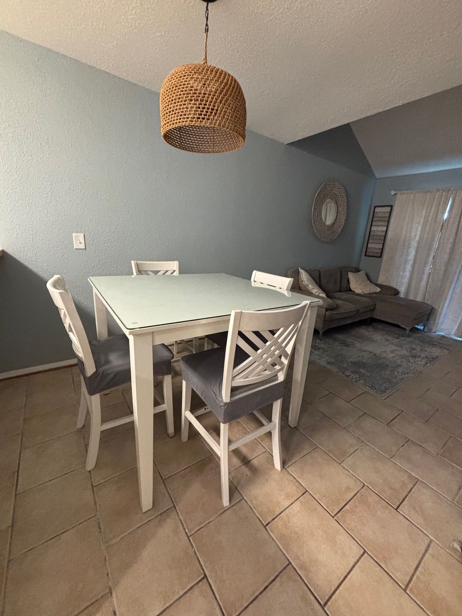 Un coin repas avec une table et des chaises blanches, un luminaire suspendu en fibres tressées, un mur bleu et un tapis à motifs.