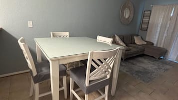 Un coin repas avec une table et des chaises blanches, un luminaire suspendu en fibres tressées, un mur bleu et un tapis à motifs.