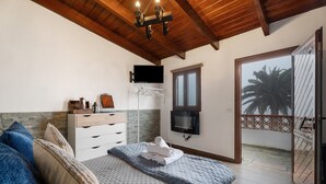 A bedroom with a bed, a dresser, a TV, a window, and a balcony with a view of palm trees.