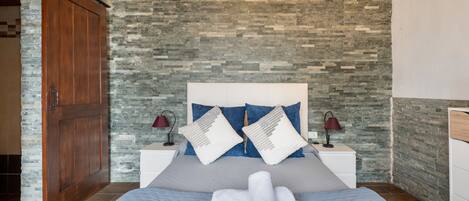A bedroom with a stone wall, a wooden door, a bed with towels, two bedside lamps, and a nightstand.
