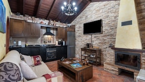 A cozy living area with a sofa, coffee table, fireplace, stone wall, wooden ceiling, and a kitchen with cabinets and appliances.