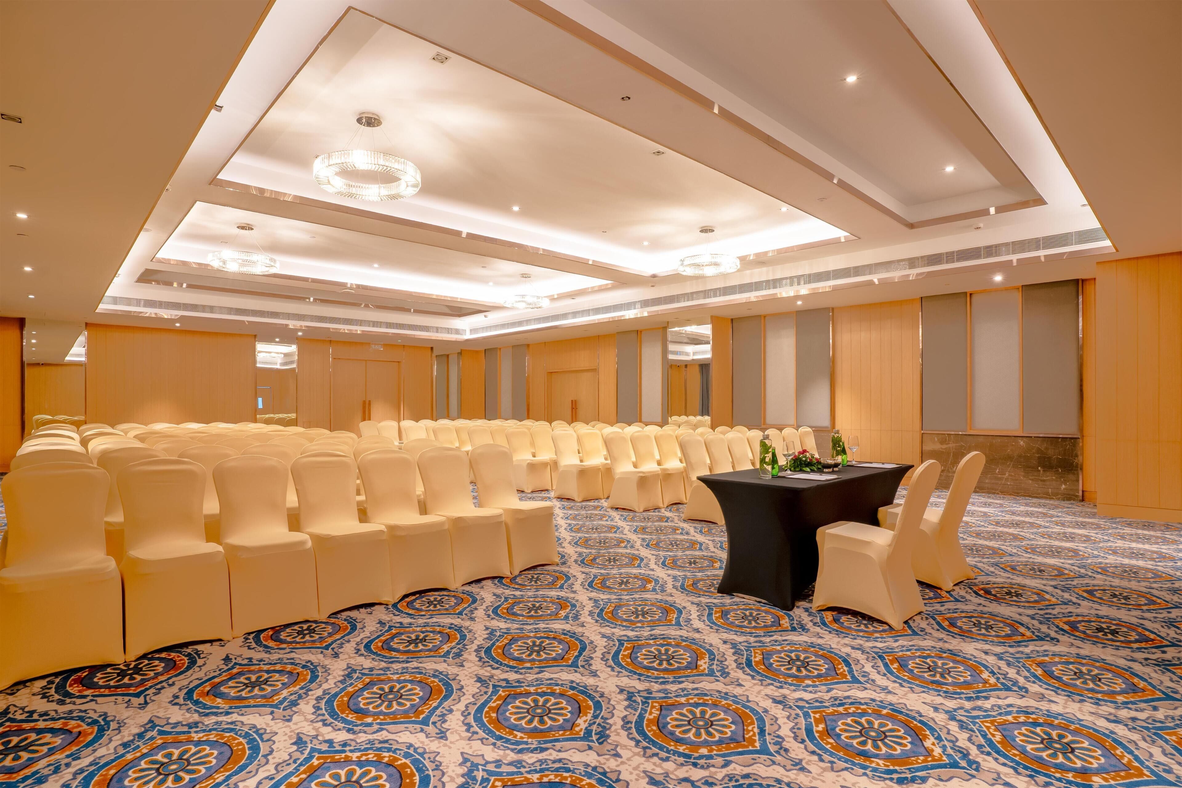 Une salle de conférence avec des rangées de chaises blanches, une table noire ornée d’un vase et un tapis à motifs.