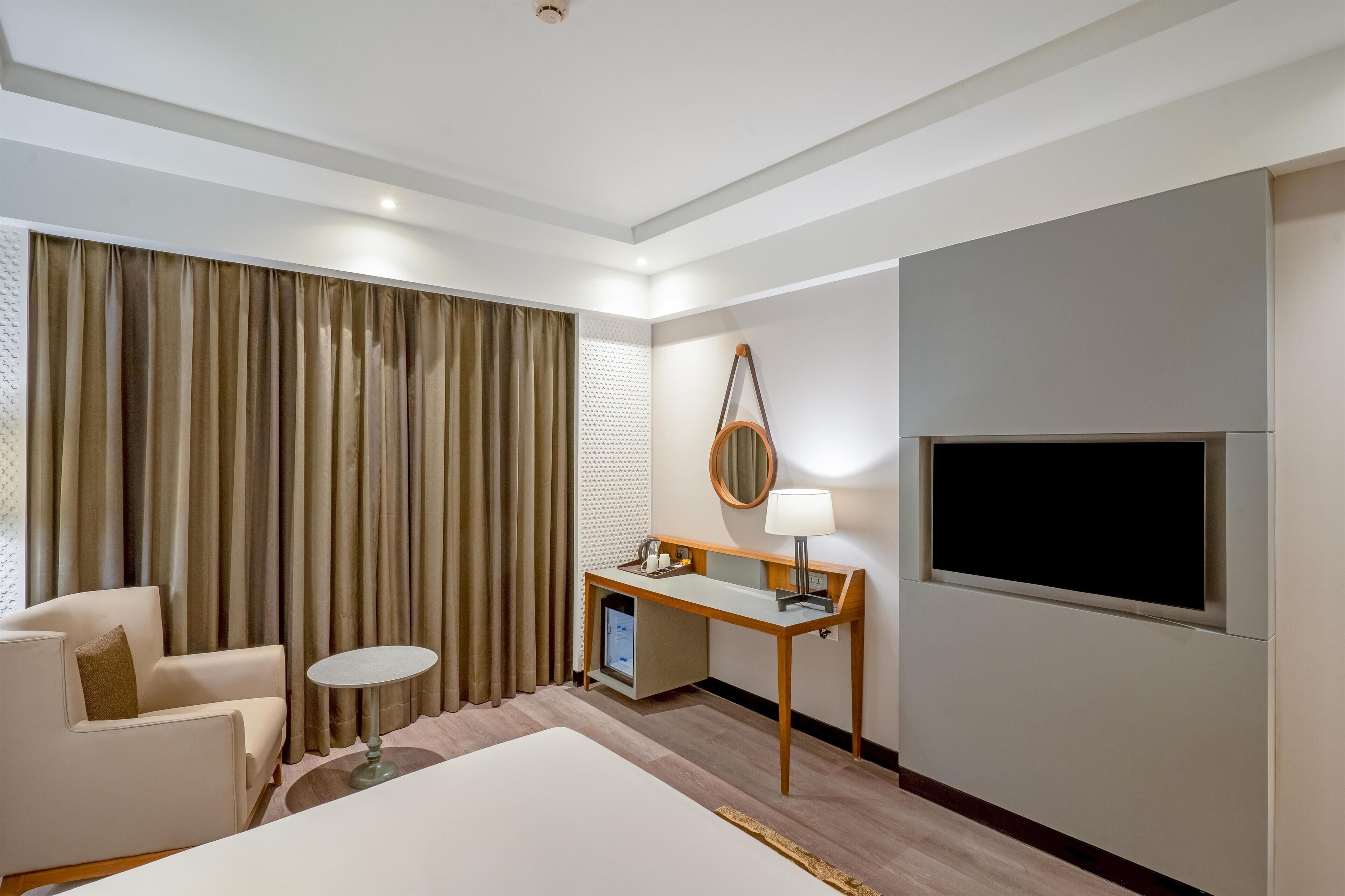 Una habitación de hotel con cama, escritorio con lámpara, televisión, silla y una ventana grande con cortinas.