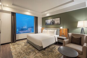 A hotel room with a large bed, a sofa, a small table, and a view of a cityscape through a large window. - Park Inn By Radisson Lucknow Vikas Nagar (Lucknow)