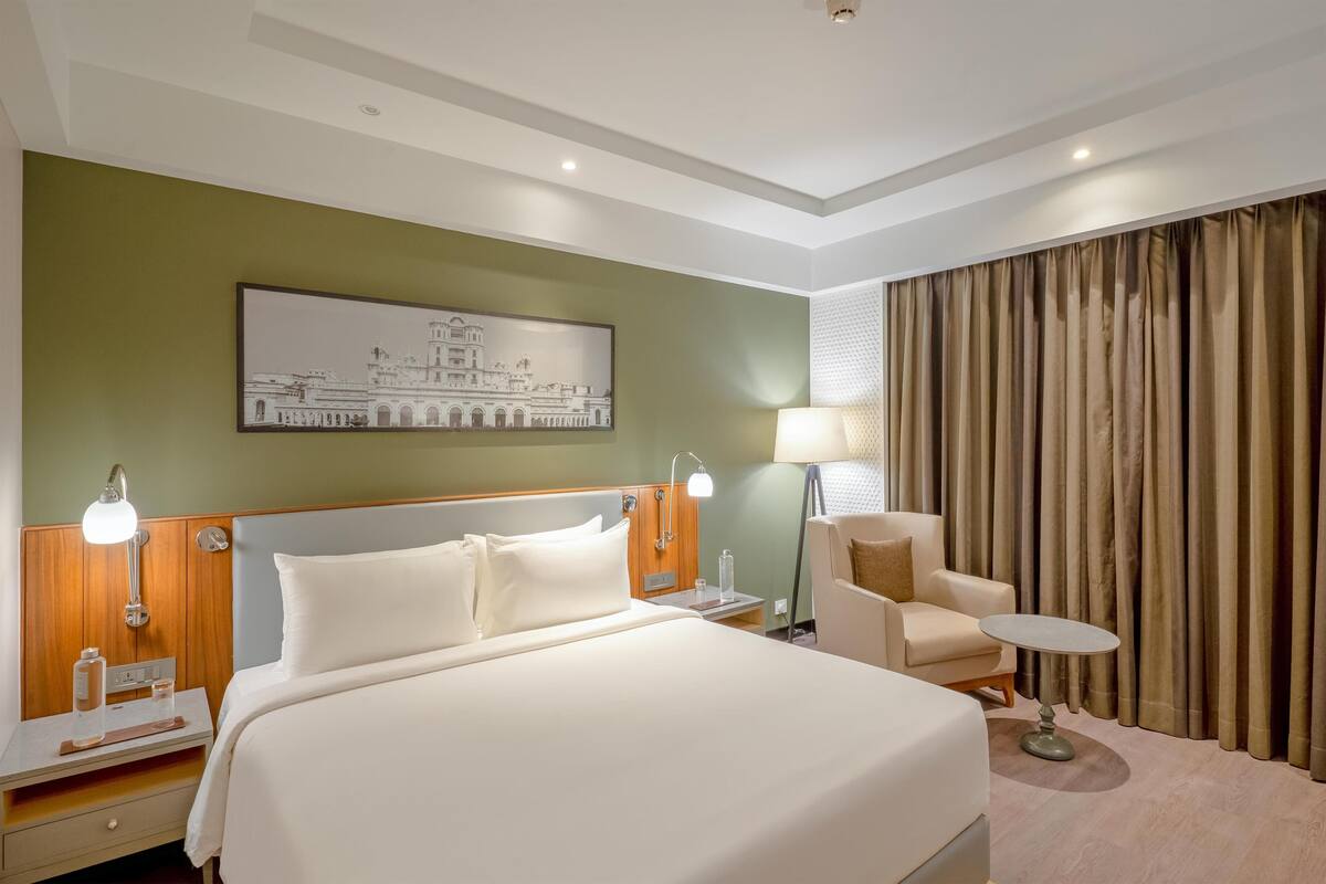 A hotel room with a large bed, a nightstand, a desk, a chair, a table, and a lamp.
