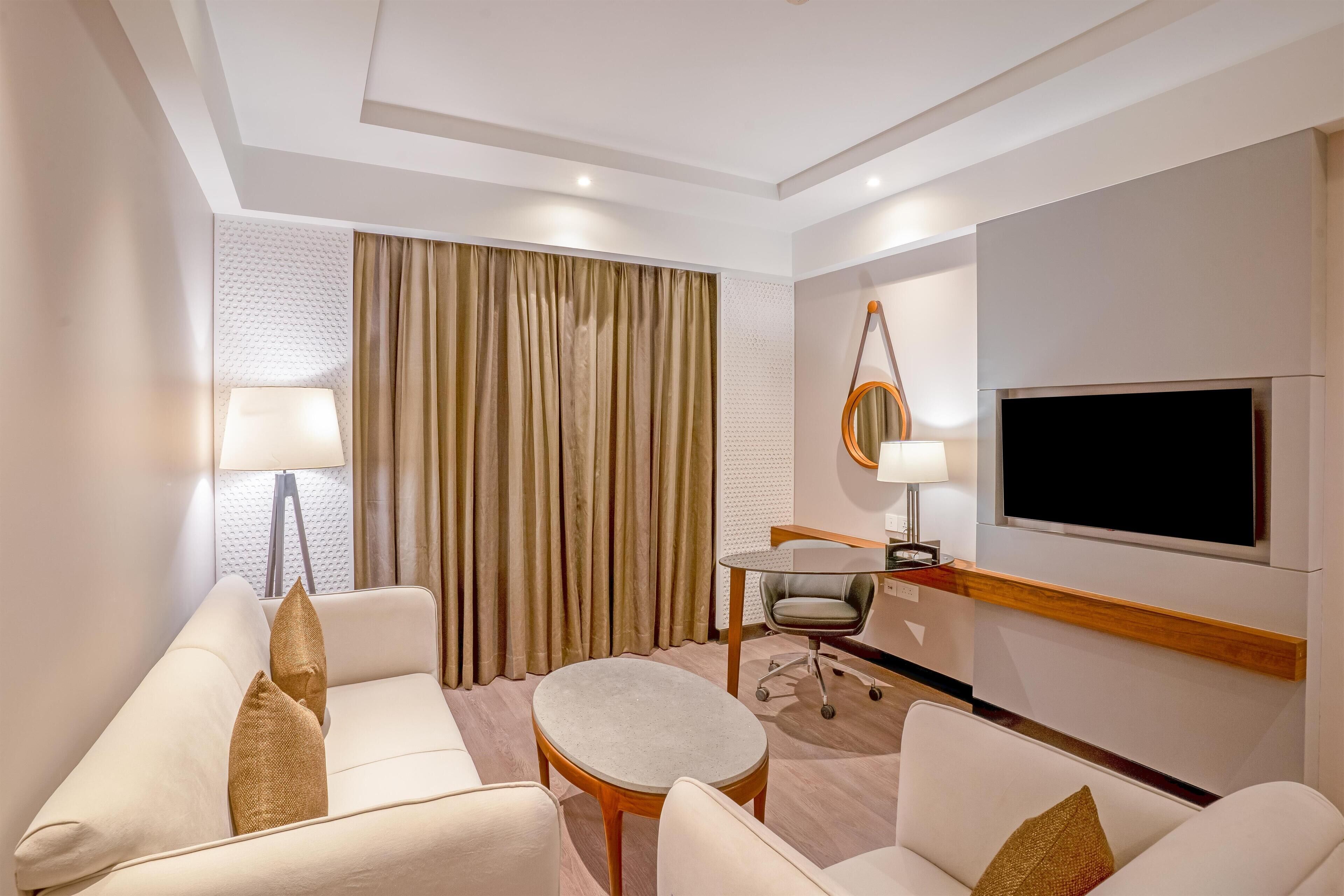 A modern hotel room with a sofa, armchair, round table, and a flat-screen TV mounted on the wall.