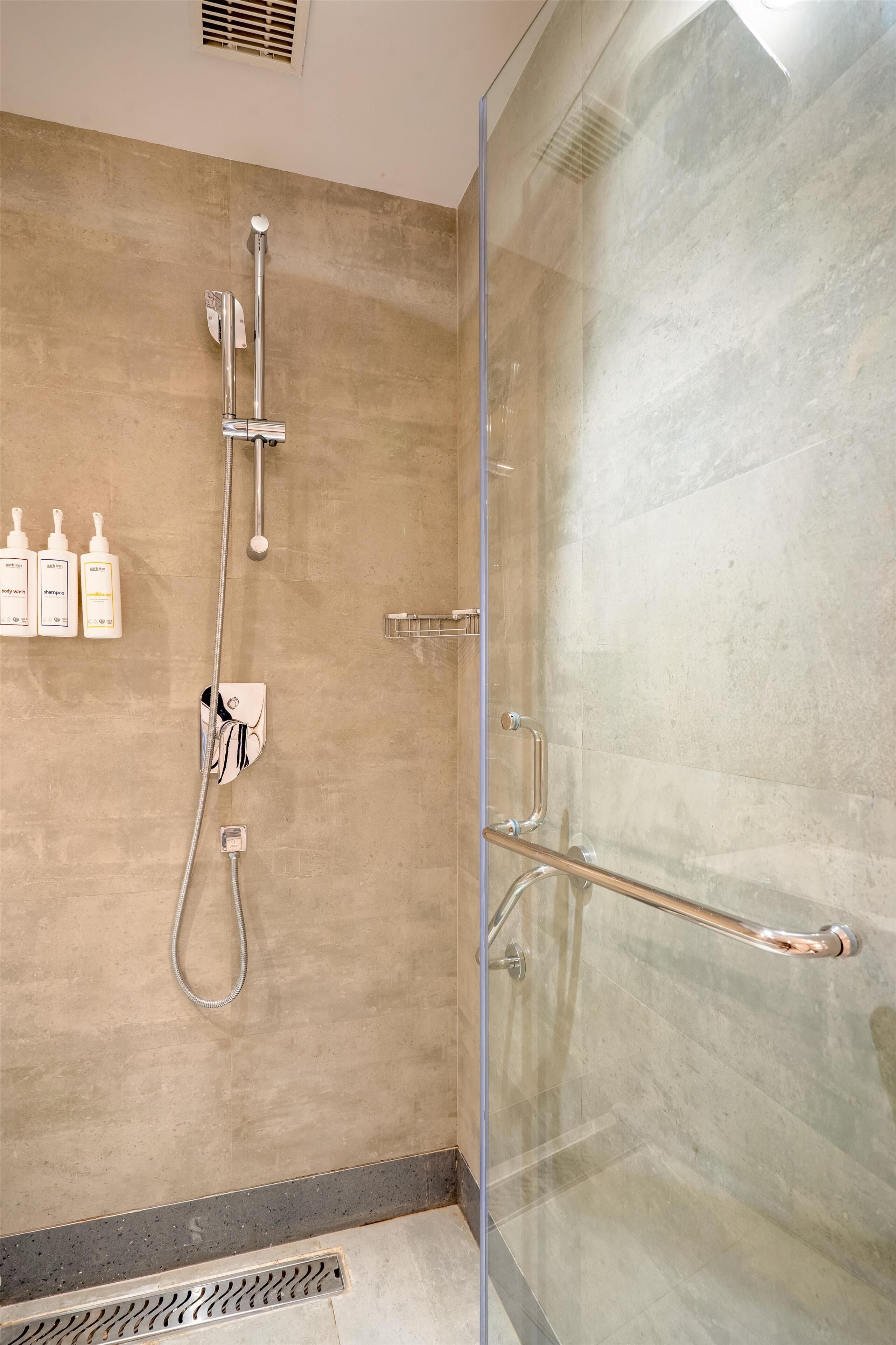 Un baño moderno con cabina de ducha de vidrio, ducha de mano y grifo empotrado. Hay tres botellas de champú y acondicionador en la pared.