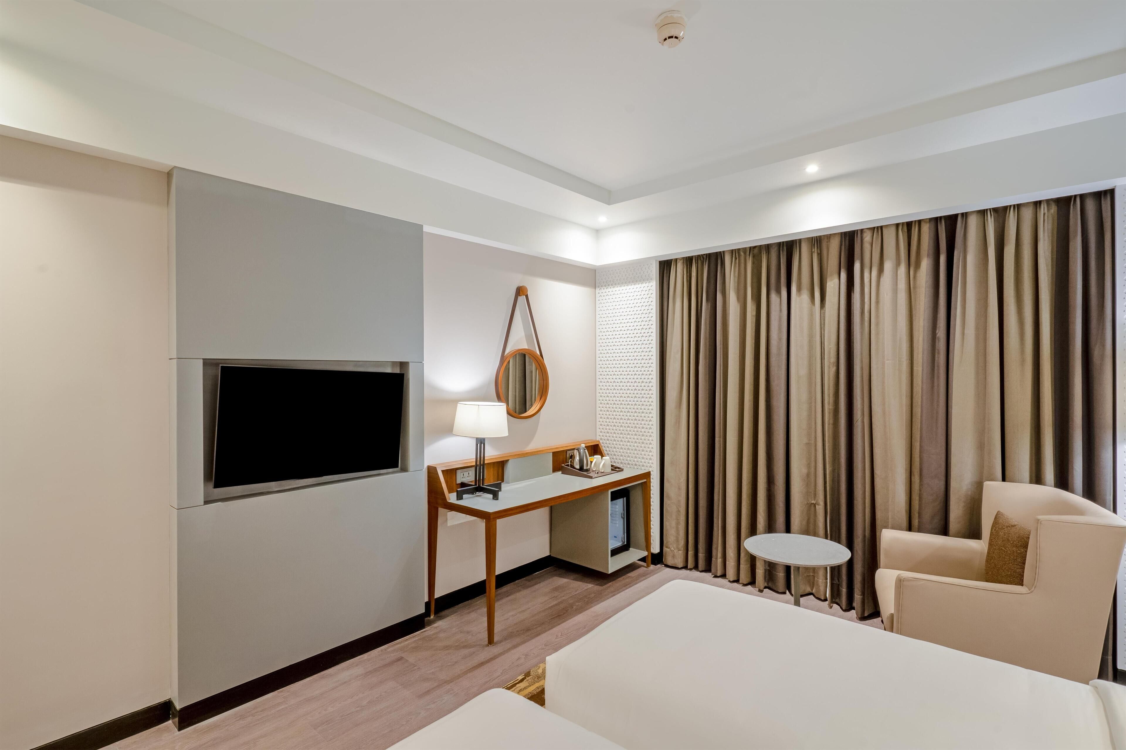 A hotel room with a bed, a desk with a lamp, a TV, a chair, and a window with curtains.