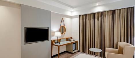 A hotel room with a bed, a desk with a lamp, a TV, a chair, and a window with curtains.
