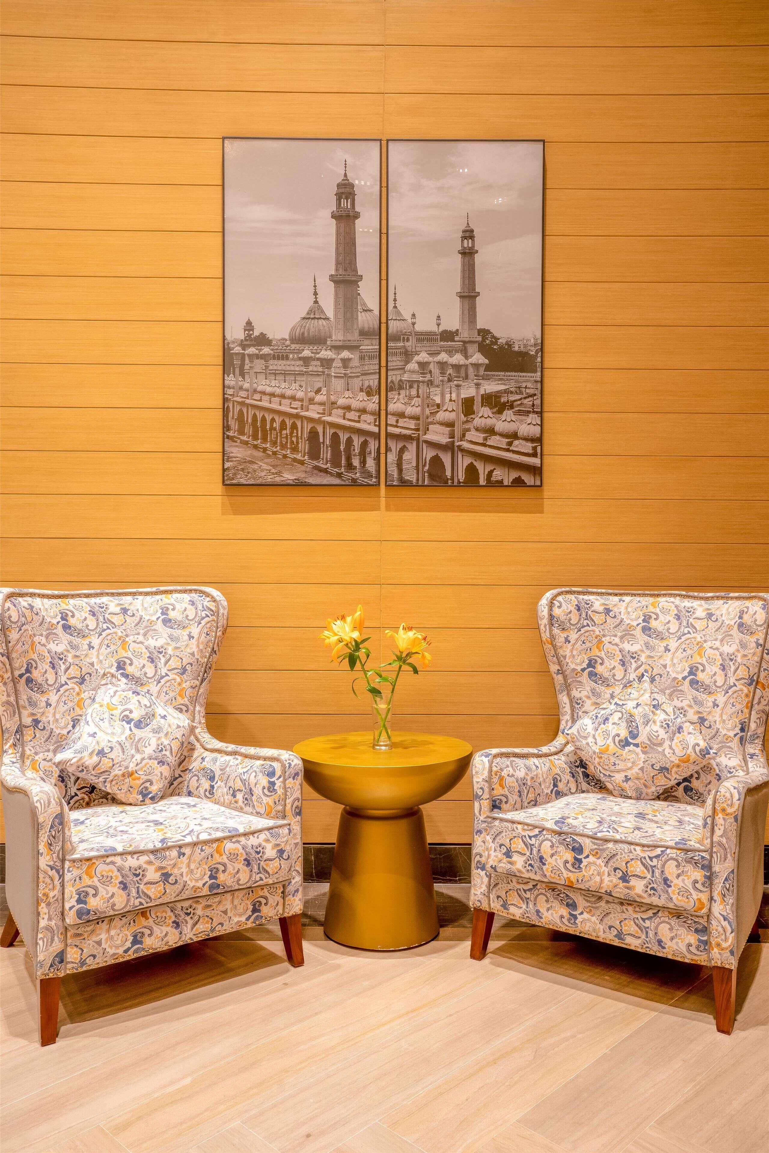 Two patterned armchairs facing each other with a small round table between them, a vase with yellow flowers, and a large framed picture of a building on the wall.