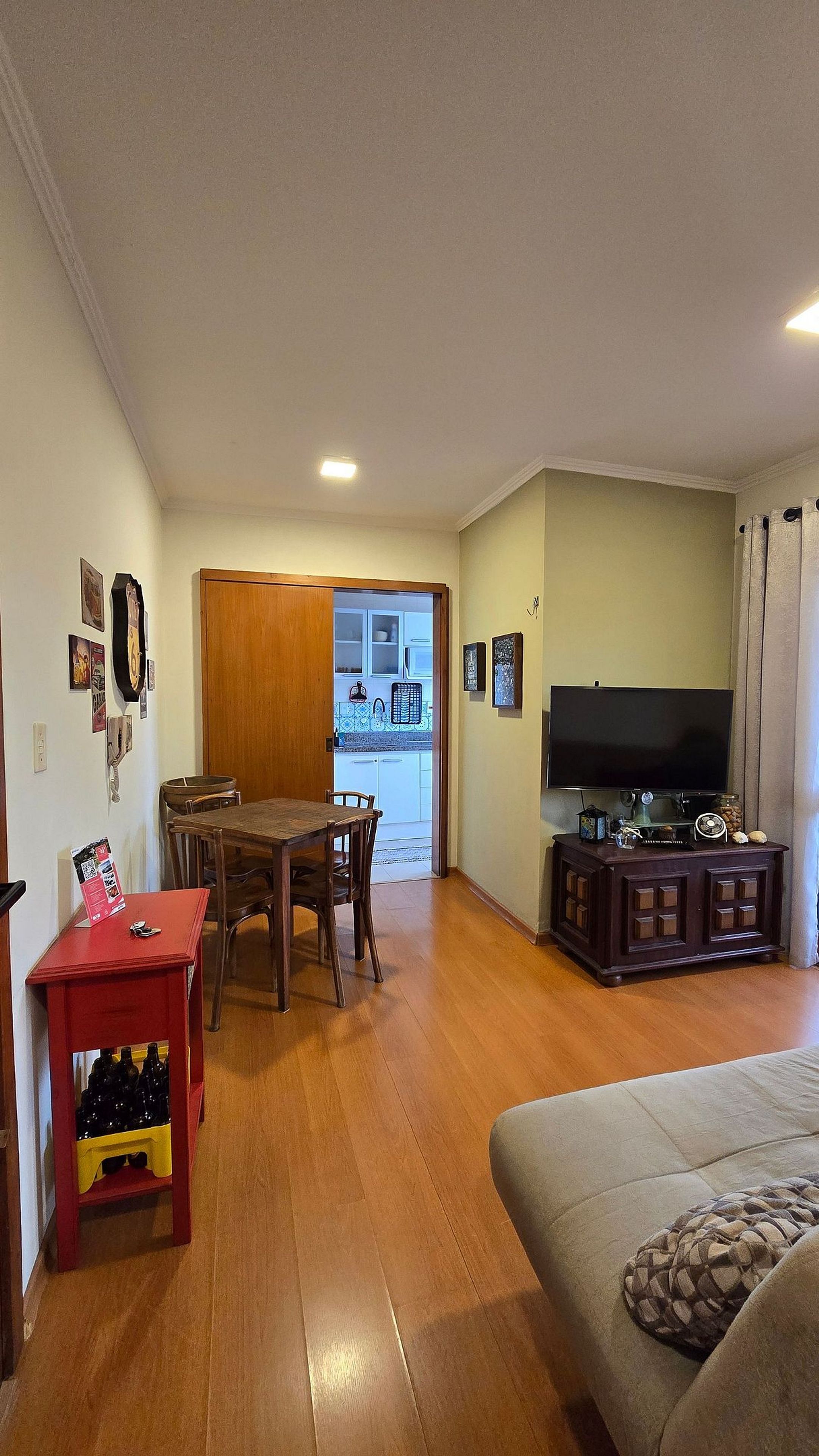 A living room with a television, a sofa, a dining table with chairs, and a kitchen visible through an open door.
