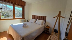 A bedroom with a large bed, a wooden headboard, a window with a view, a bedside table, and a wooden drying rack.