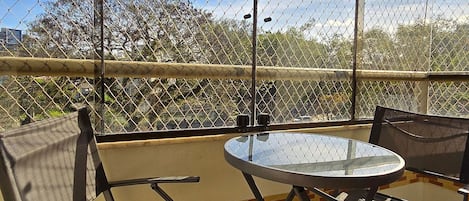 A covered outdoor seating area with a glass table and metal chairs, overlooking a fenced area with trees.