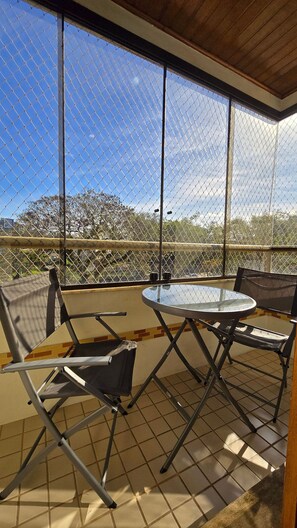 A covered outdoor seating area with a glass table and metal chairs, overlooking a fenced area with trees.