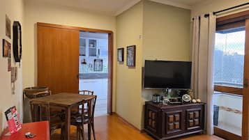 Sala de estar com mesa de jantar de madeira, cadeiras, televisão sobre um móvel e janela com cortinas.