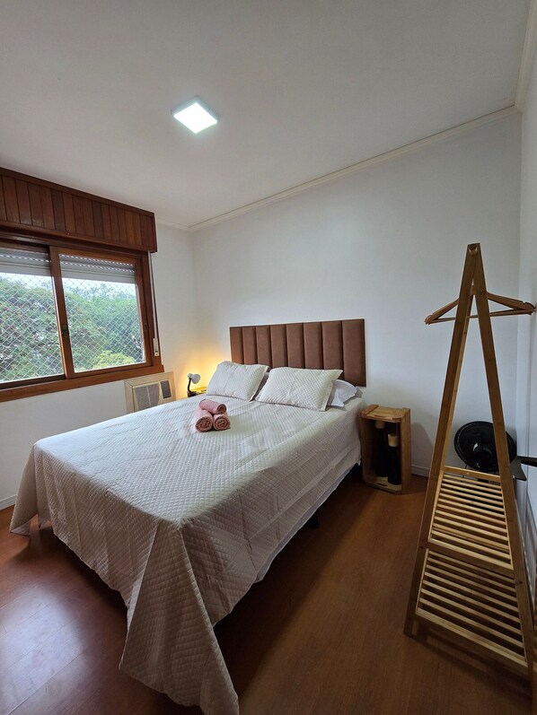 A neatly made bed with a headboard, a bedside table with a lamp, a wooden chair, and a window with a view of greenery.