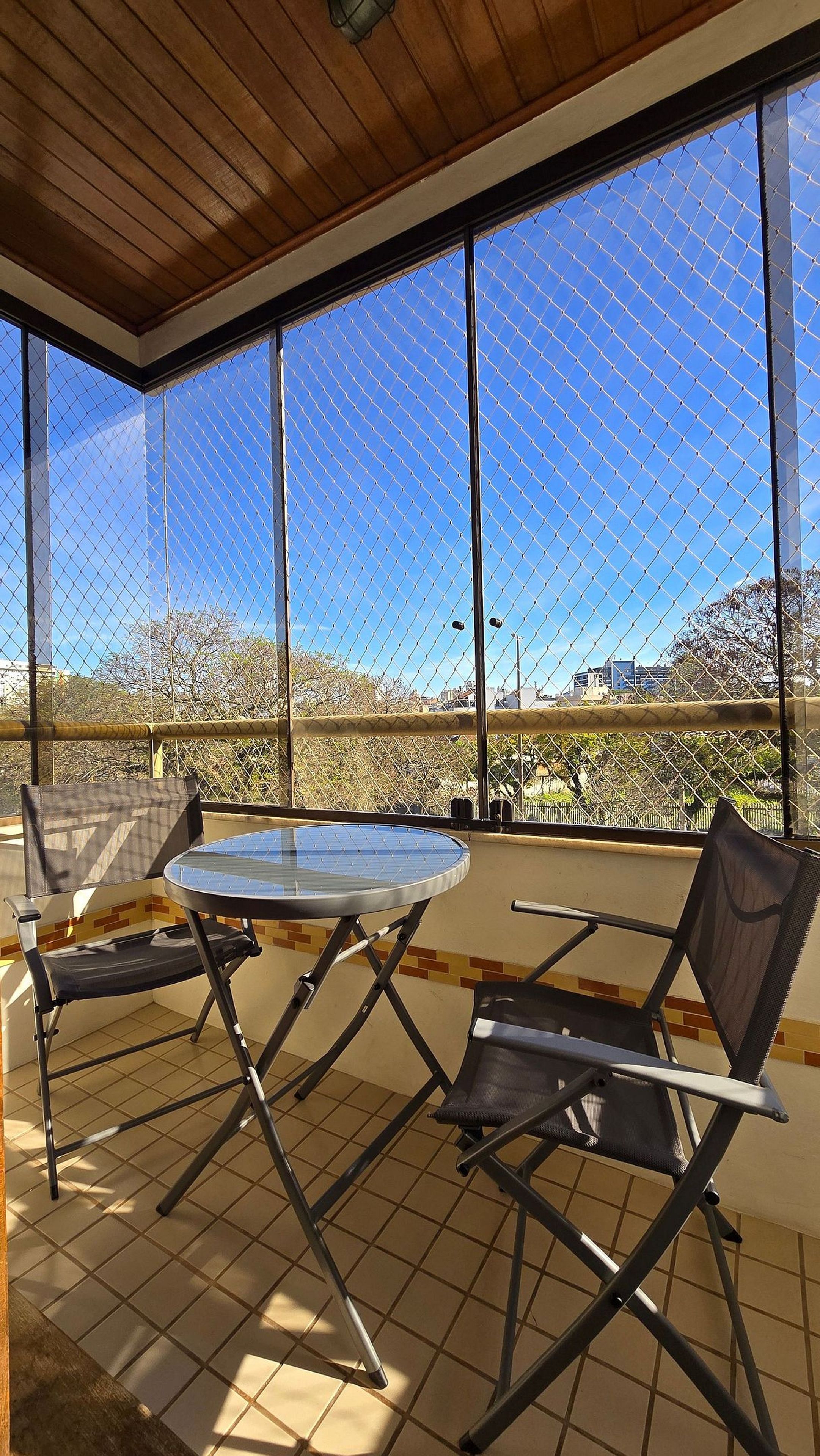 Uma área de varanda coberta com mesa, cadeiras, janelas amplas e vista para os arredores.