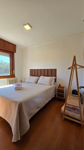 A bedroom with a bed, a bedside table, a wooden hanger, and a window.