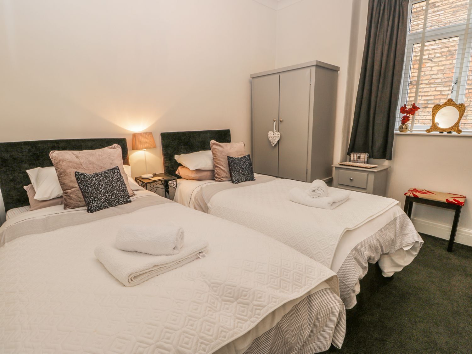 A hotel room with two beds, a wardrobe, a nightstand with a lamp, and a window with curtains.