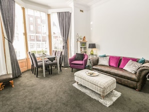 A living room with a dining area, a purple armchair, a brown leather sofa with colorful cushions, and a white ottoman.