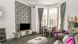 A modern living room with a flat-screen TV, a dining area with a table and chairs, and a cozy seating area with a sofa and ottoman. - Esplanade Gardens, Apartment 1 (Scarborough)