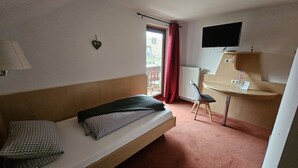 A single bed with a checkered pillow, a bedside table with a lamp, a chair, a TV mounted on a wall, and a window with a red curtain. - Hotel Restaurant Ampertal (Haag an der Amper)