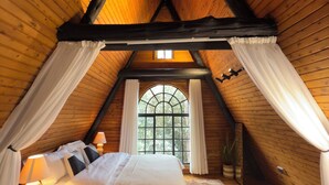 Un dormitorio acogedor en el ático con techo de madera, un ventanal con cortinas, una cama con sábanas blancas y una mesita de noche pequeña.
