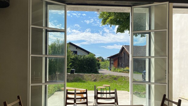 A dining area with a table and chairs, large windows offering a view of a rural landscape with houses and greenery. - Landsberg artist farmhouse - Unique (Pürgen)