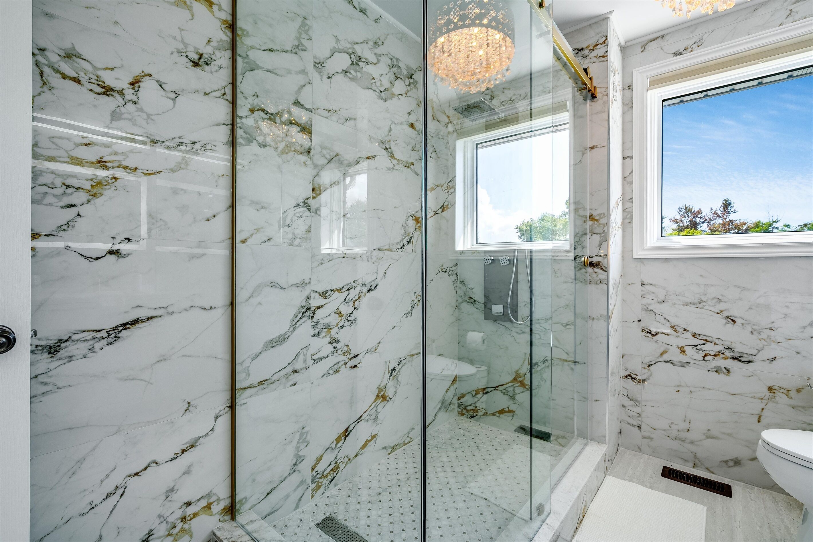 Une salle de bain aux murs recouverts de marbre, une douche en verre, une fenêtre offrant une vue agréable et un lustre.