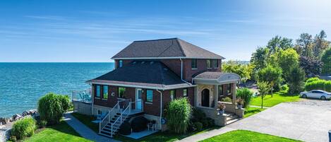 A two-story house with a balcony overlooking the sea, a driveway, and a lawn with outdoor furniture.
