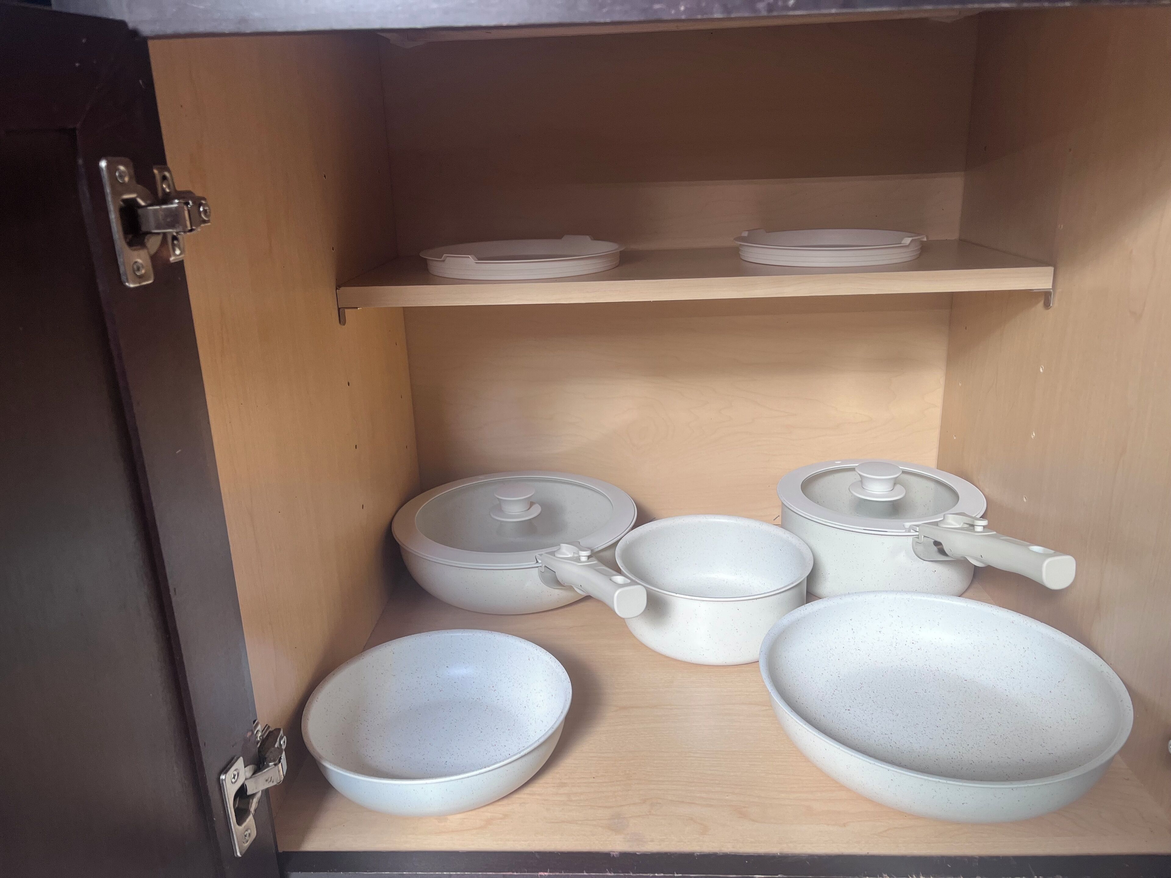 A kitchen cabinet with white ceramic cookware, including pots, pans, and bowls.