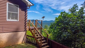 A wooden cabin with a wooden staircase leading to a deck, surrounded by lush greenery and mountains in the background. - Whisper Mountain Retreat (Franklin)