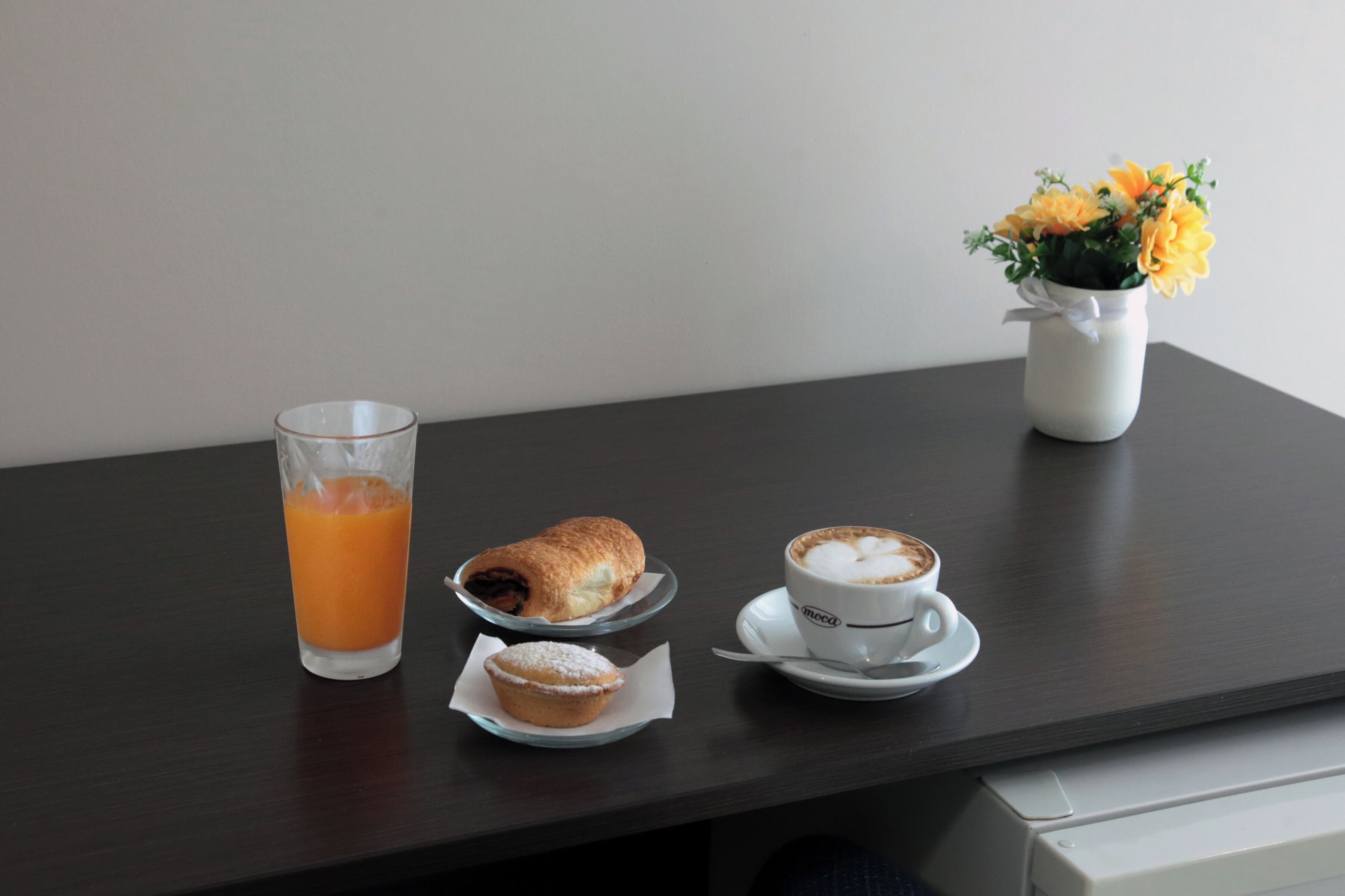 Un tavolo con un bicchiere di succo d'arancia, un pasticcino, un muffin e una tazza di caffè, con un vaso di fiori sullo sfondo.