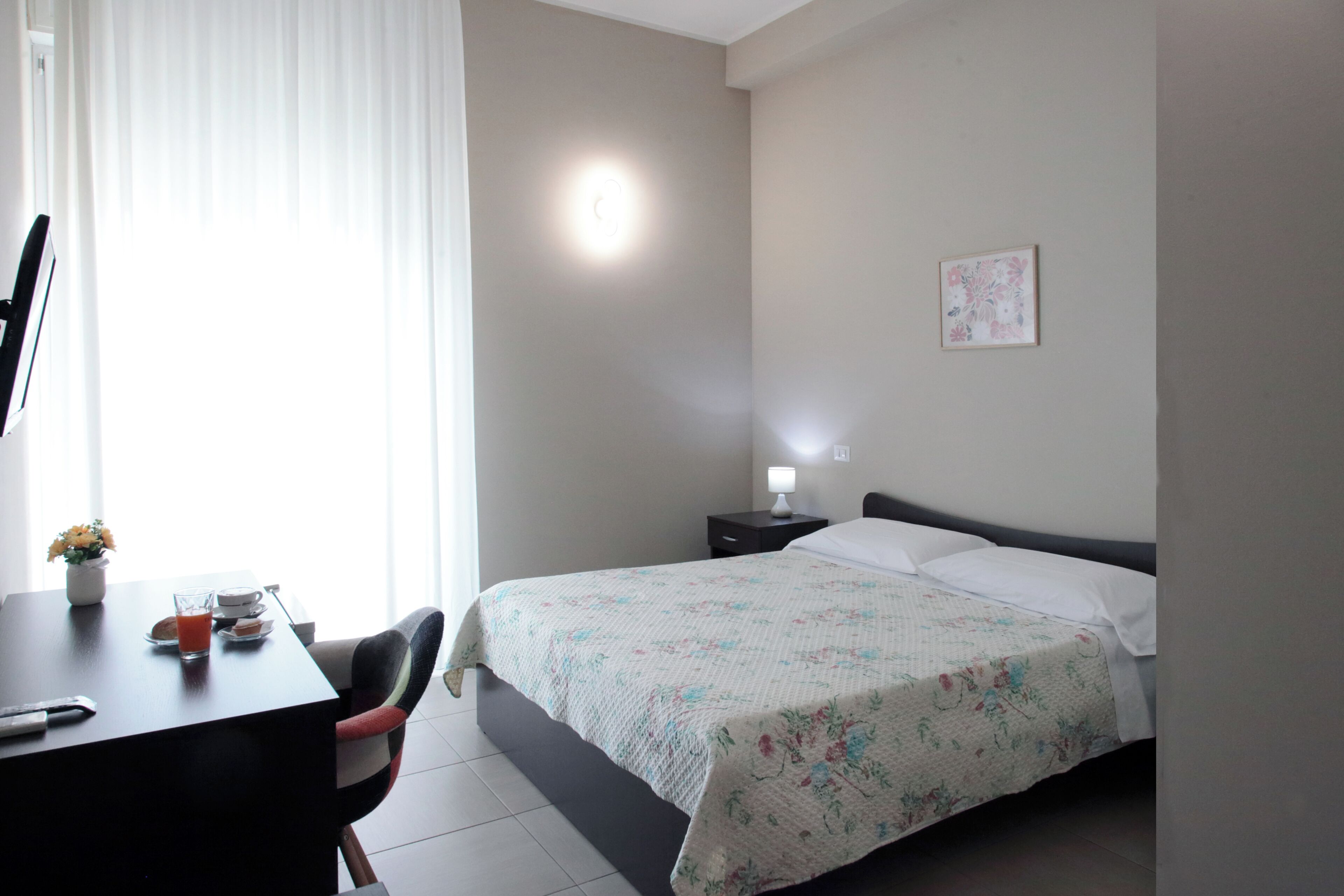 A hotel room with a bed, a desk with a chair, a nightstand with a lamp, and a window with curtains.