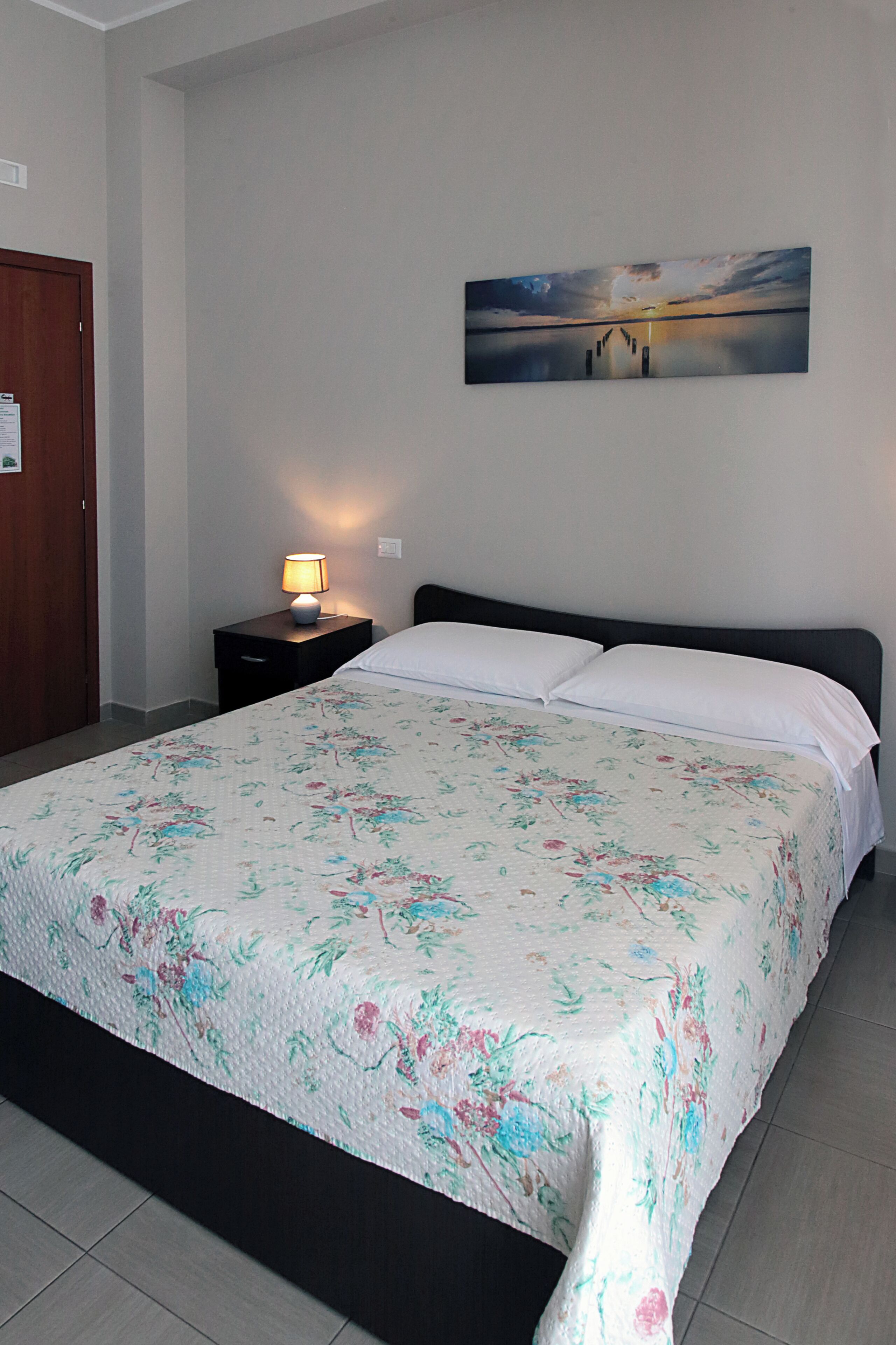 A bedroom with a bed, a nightstand with a lamp, a painting on the wall, and a wooden door.