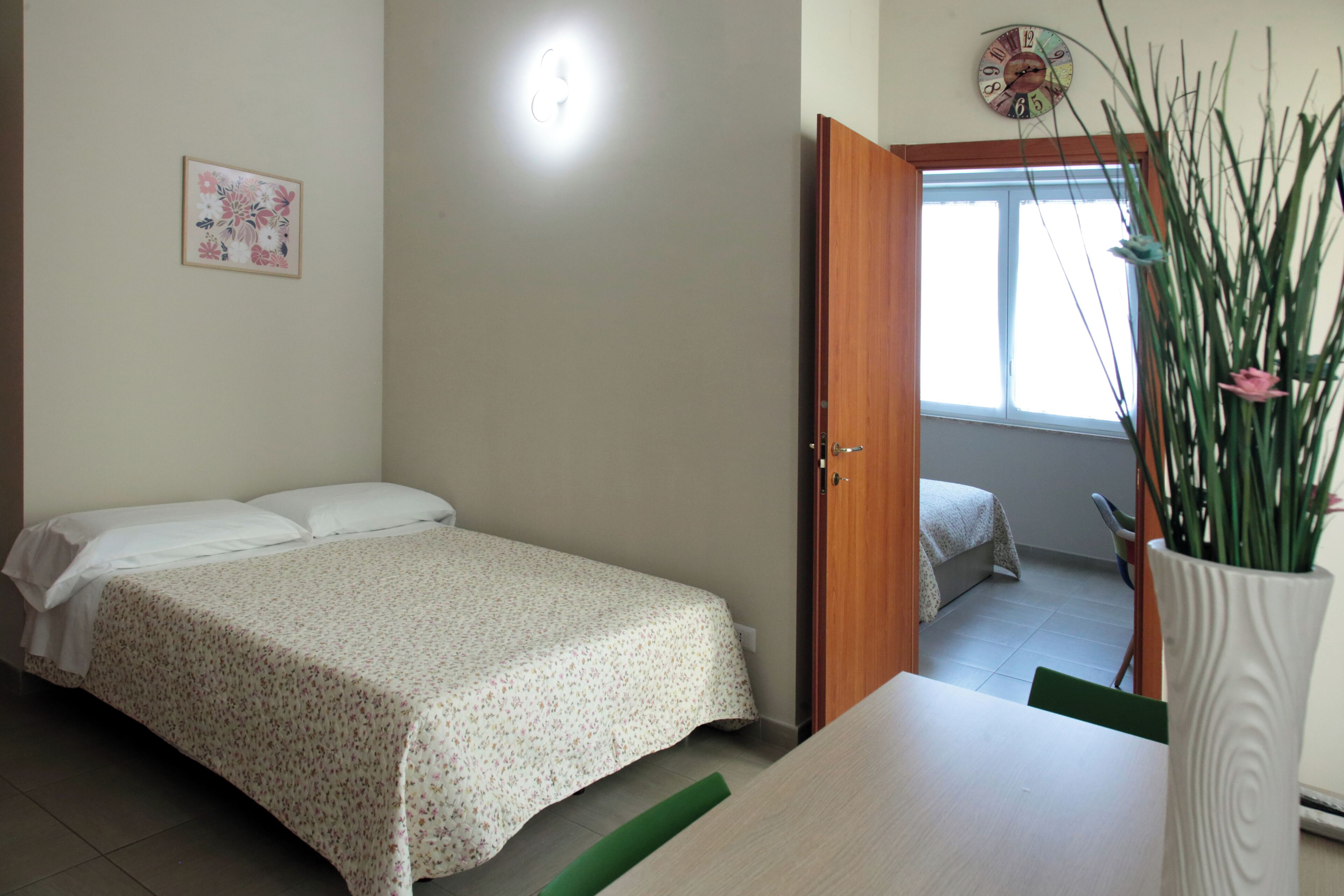A bedroom with a bed, a bedside table, a green mat, a wall clock, and a window with a view of the outside.