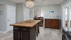 Une cuisine dotée d’un îlot central en bois, de meubles de rangement sombres, d’un lustre et d’un coin repas avec des chaises et une table.