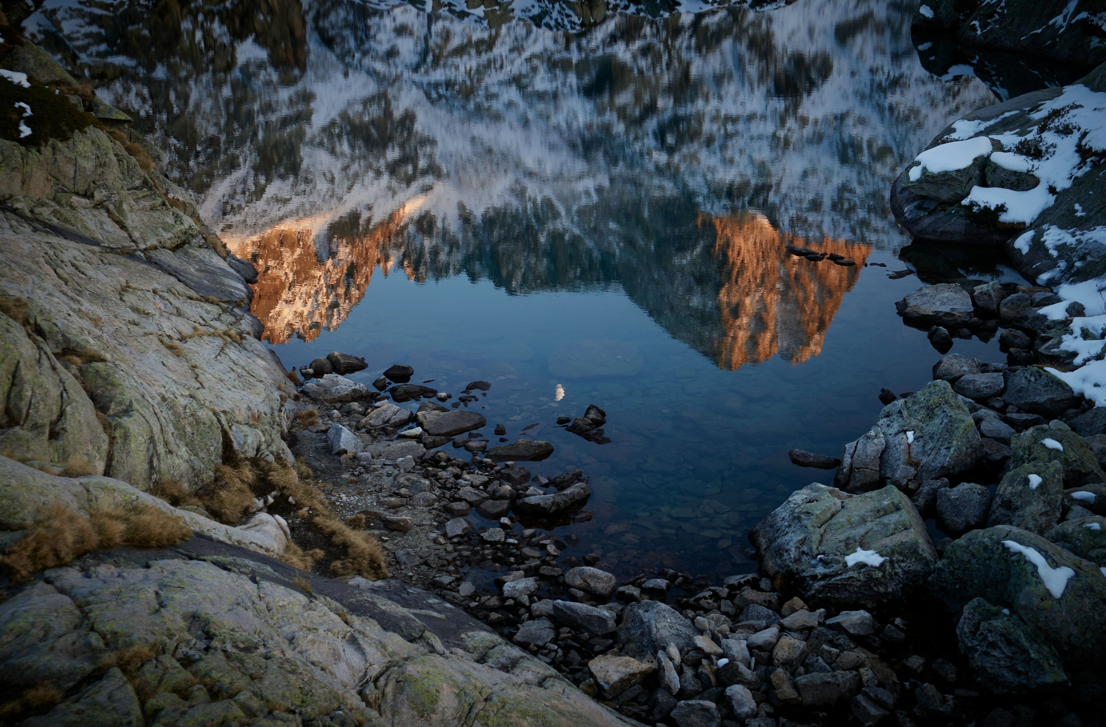 Una costa rocosa con un cuerpo de agua tranquilo que refleja árboles y una puesta de sol.