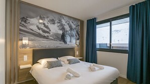 Habitación de hotel con una cama grande, cabecera con pintura de un paisaje de montaña, ventana con cortinas y vistas a un paisaje nevado.