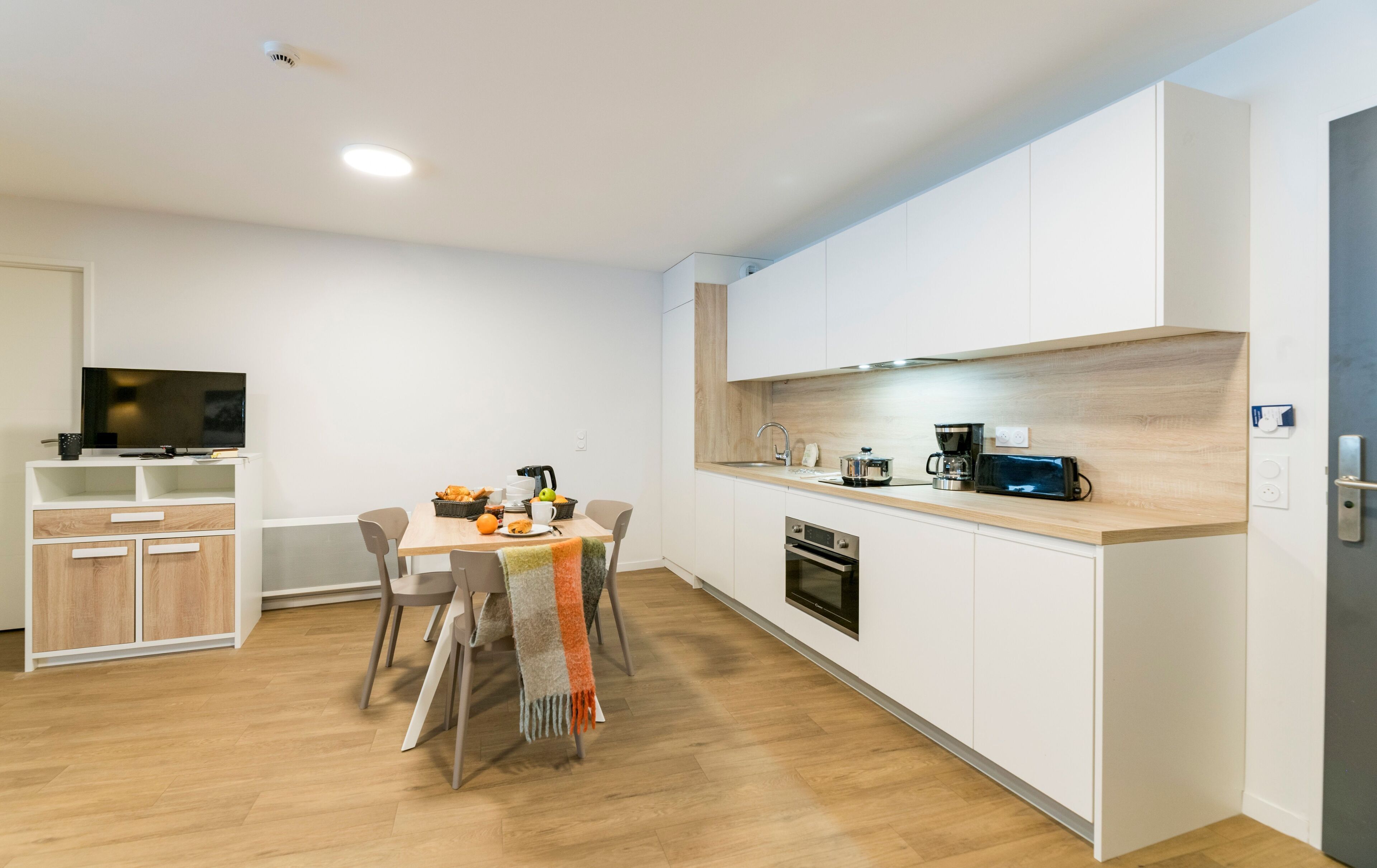 A modern kitchen with a dining area, wooden cabinets, and a TV.