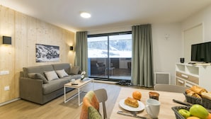 A modern living room with a grey sofa, a coffee table, and a dining area with a table set for breakfast. A large window offers a view of a snowy landscape.