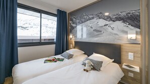 A hotel room with two beds, a large window with curtains, a wall-mounted painting of a snowy mountain, and a bedside table with a lamp.