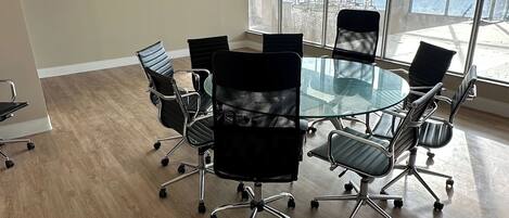 A conference room with a glass table and black office chairs, large windows, and wooden flooring.