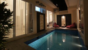 A modern indoor pool area with a clear glass wall, a small seating area with cushions, and a decorative light fixture. - Azura Villas Nusa Dua Bali (Nusa Dua)