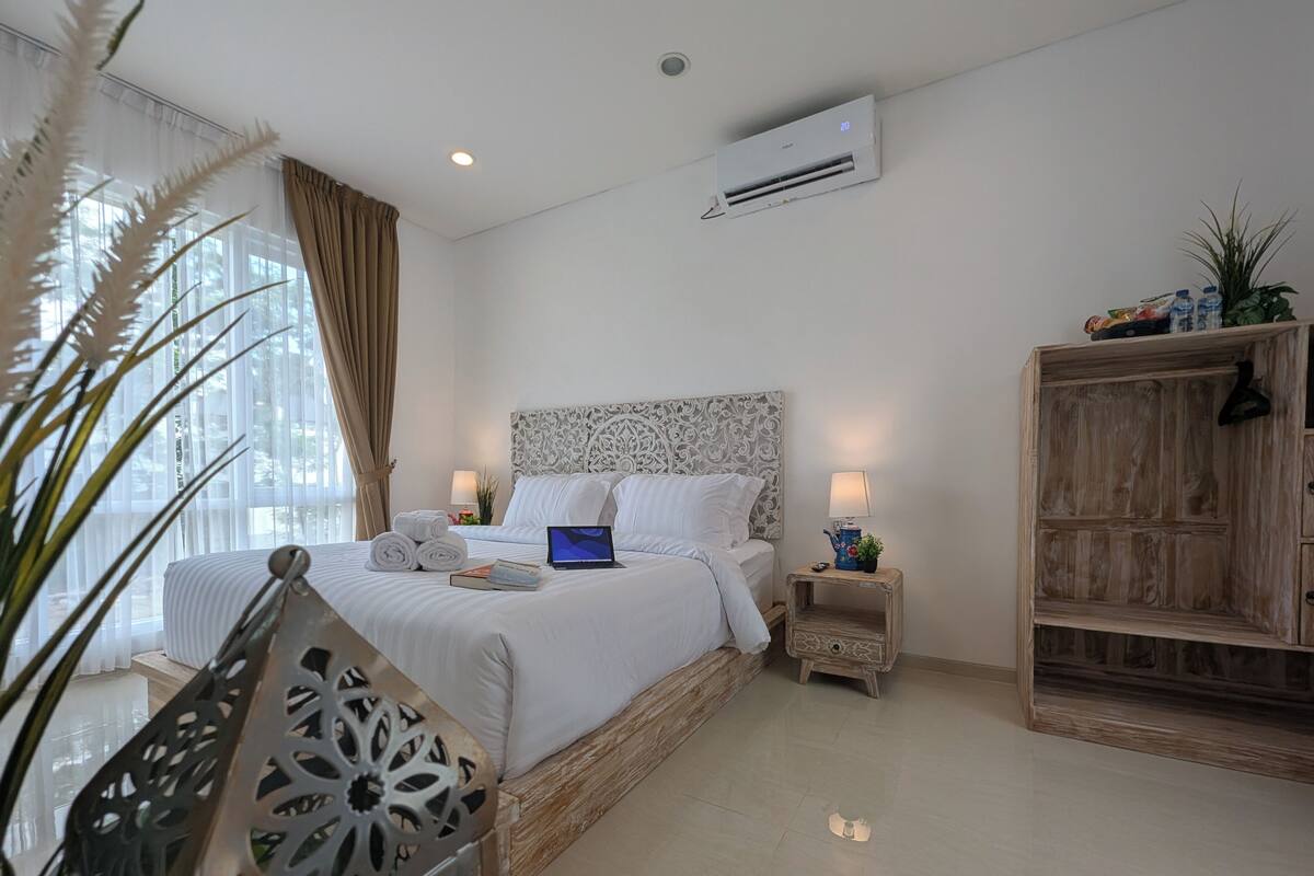 A hotel room with a bed, air conditioning unit, bedside lamp, laptop, and a wooden cabinet.