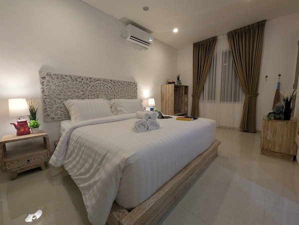 A neatly made bed with a headboard, a bedside table with a lamp, a small cabinet, and a potted plant. - Azura Villas Nusa Dua Bali (Nusa Dua)