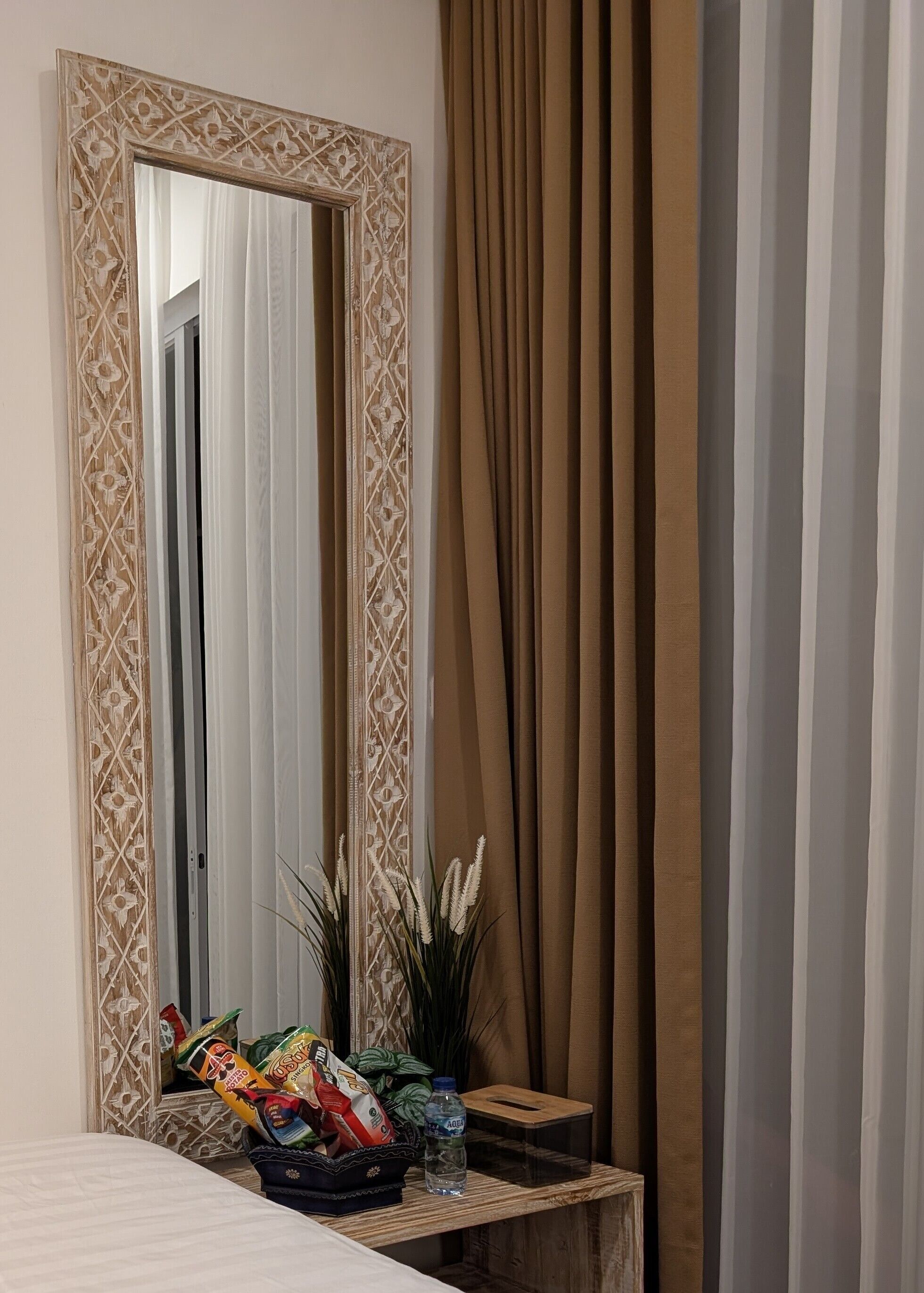 A room with a large mirror, a bedside table with snacks, a bottle, and a plant.
