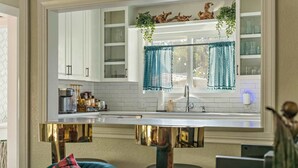 A modern kitchen with white cabinets, a stainless steel countertop, and a gold base. There is a window with teal curtains and a hanging plant. - Poolside Funhouse with Arcade, Billiard, Poker (Austin)