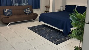 Quarto com cama, sofá, ventoinha no teto, janela com cortinas, abajur e parede com duas molduras.