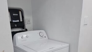 A compact, white front-loading washer and dryer unit in a corner of a room with a tiled floor and a small potted plant. - The Castle ᴶ (Philipsburg)